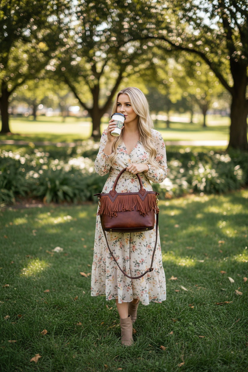 Woman in a floral dress holding a coffee cup and brown handbag in a park.
