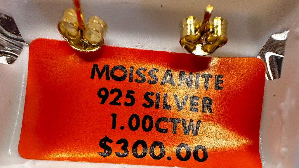 Gold earrings with a label and a coin for scale, on a reflective surface.