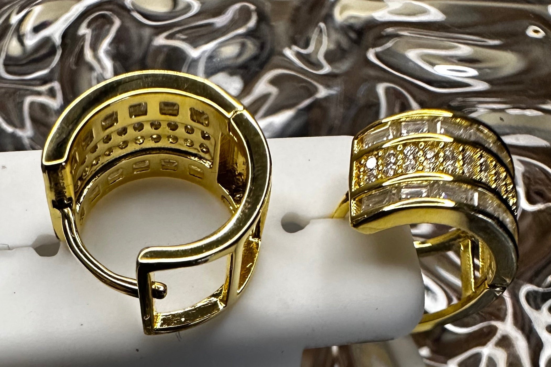 Gold hoop earrings on a white stand with a quarter coin in the center, against a reflective metallic background.