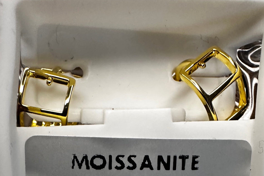 Gold moissanite earrings in a display case with a coin for scale on a reflective surface.