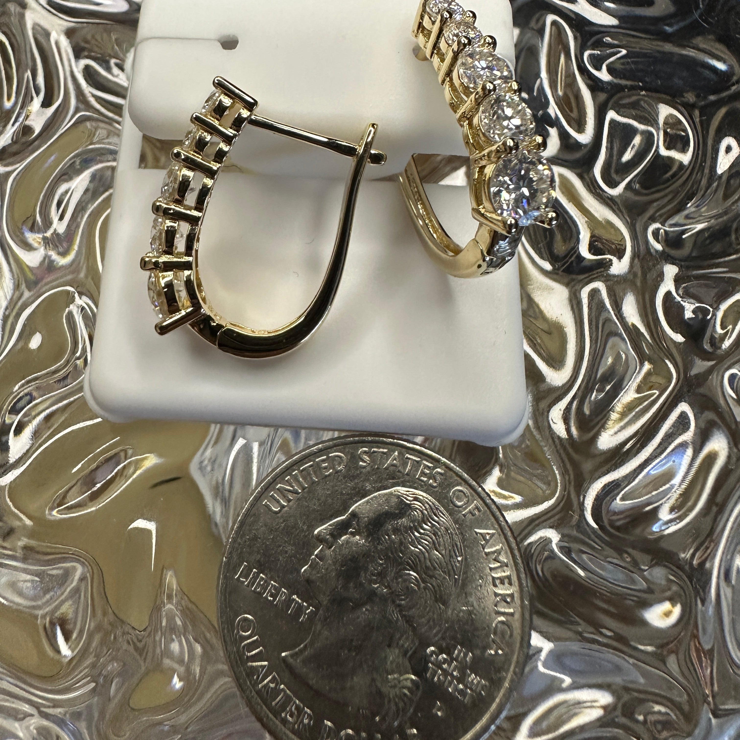 Gold hoop earrings on a white stand with a quarter for scale, on a reflective surface.