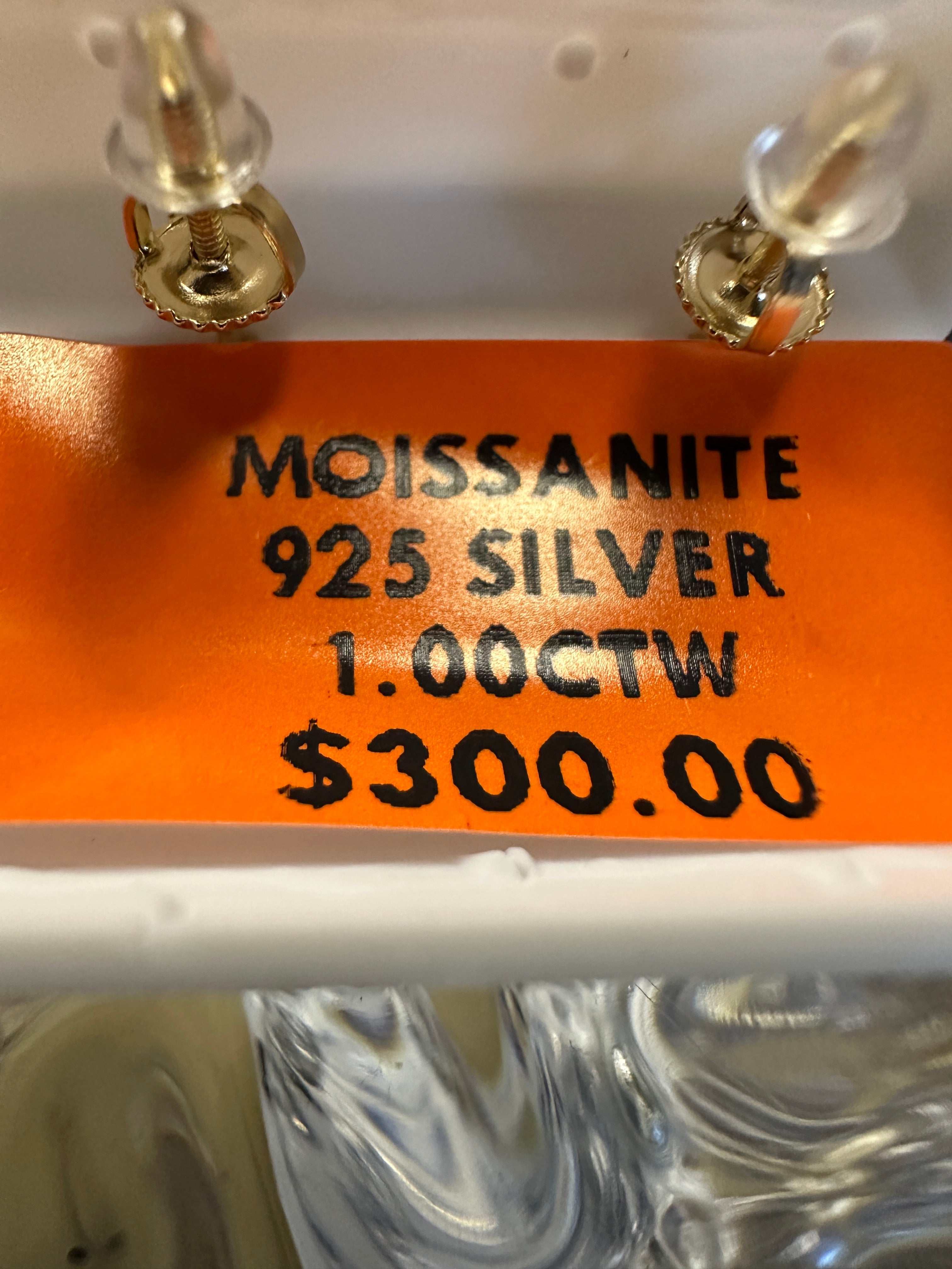Earring with a label indicating 'Moissanite 925 Silver 1.00CTW $300.00' on an orange background.