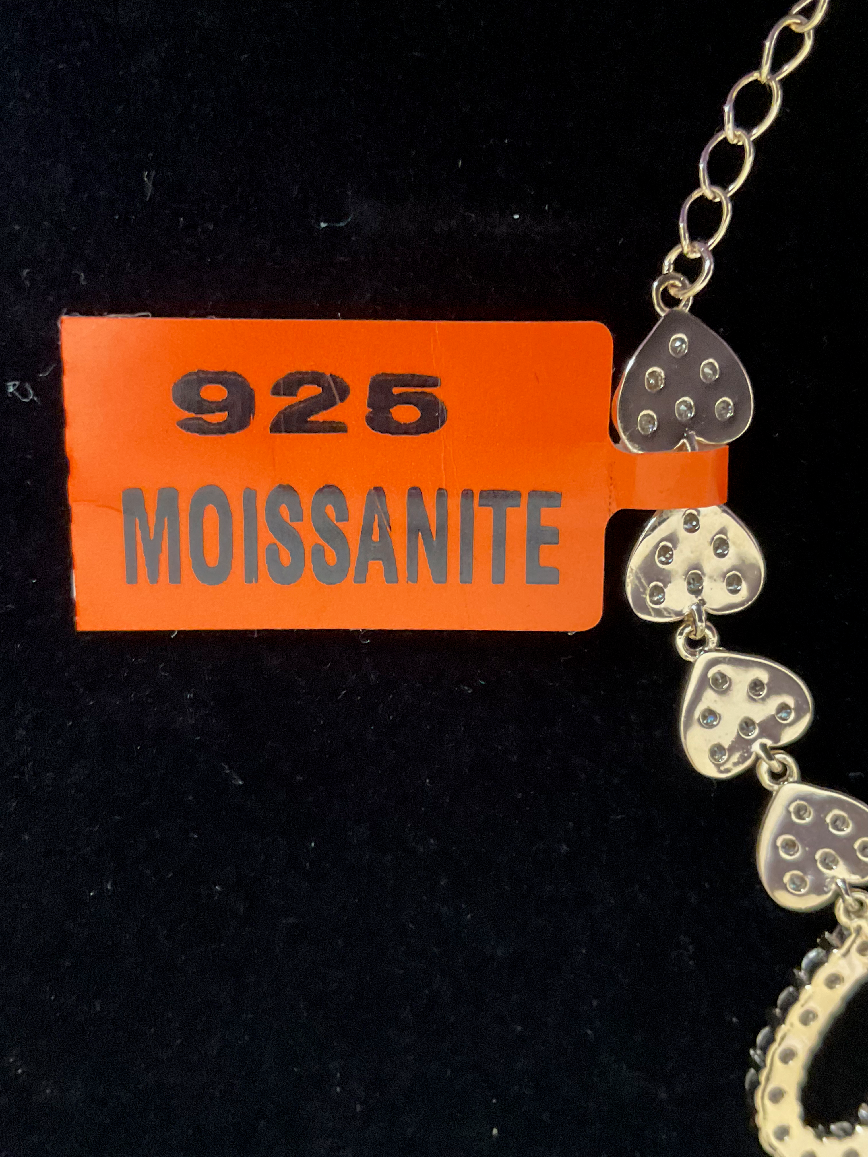 Gold bracelet with heart-shaped charms on a black background, featuring an orange tag with '925 Moissanite' text.
