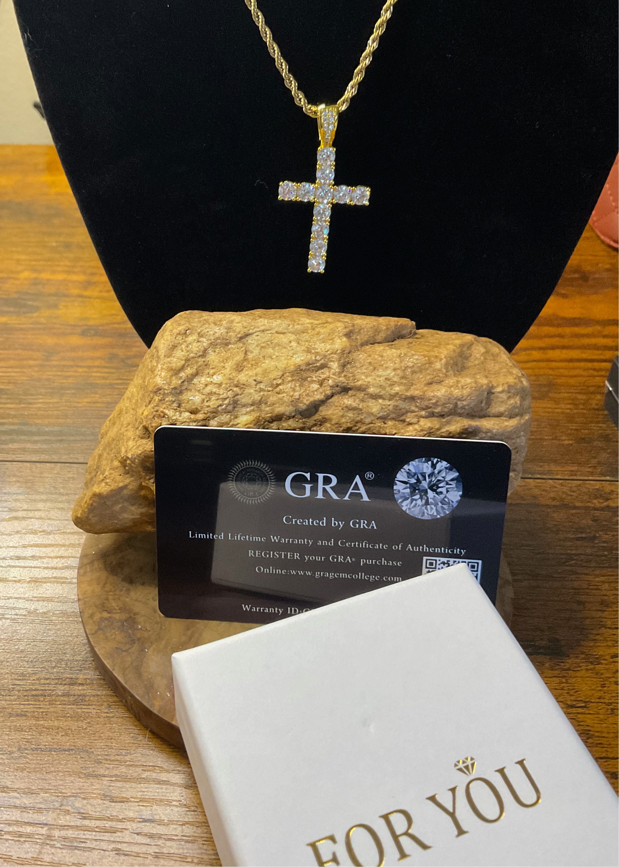 Gold cross necklace on a stand with a GRA card and 'For You' box on a wooden surface