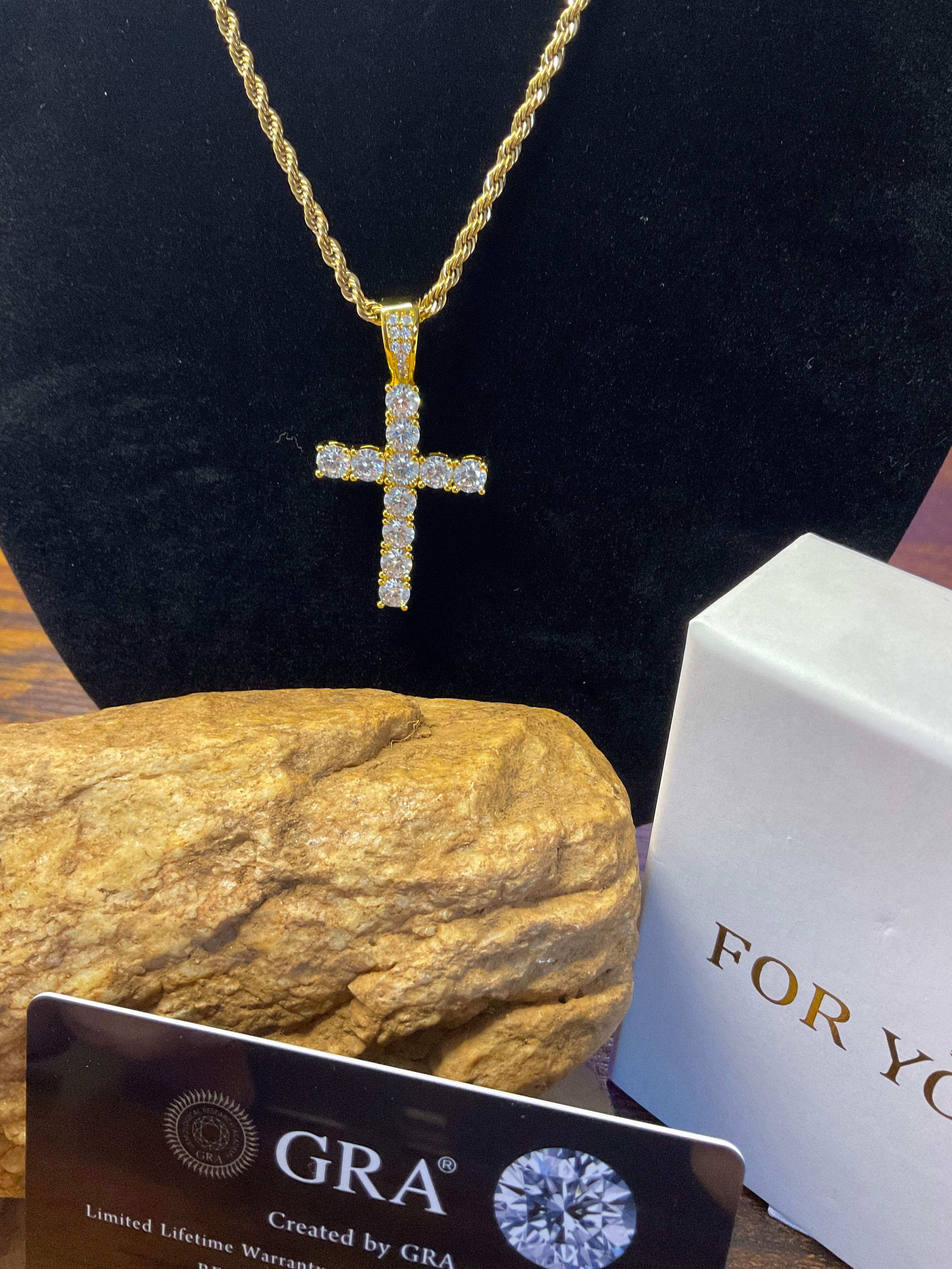 Gold cross necklace with diamond accents on a black stand, next to a rock and a white box with 'GRA' branding.