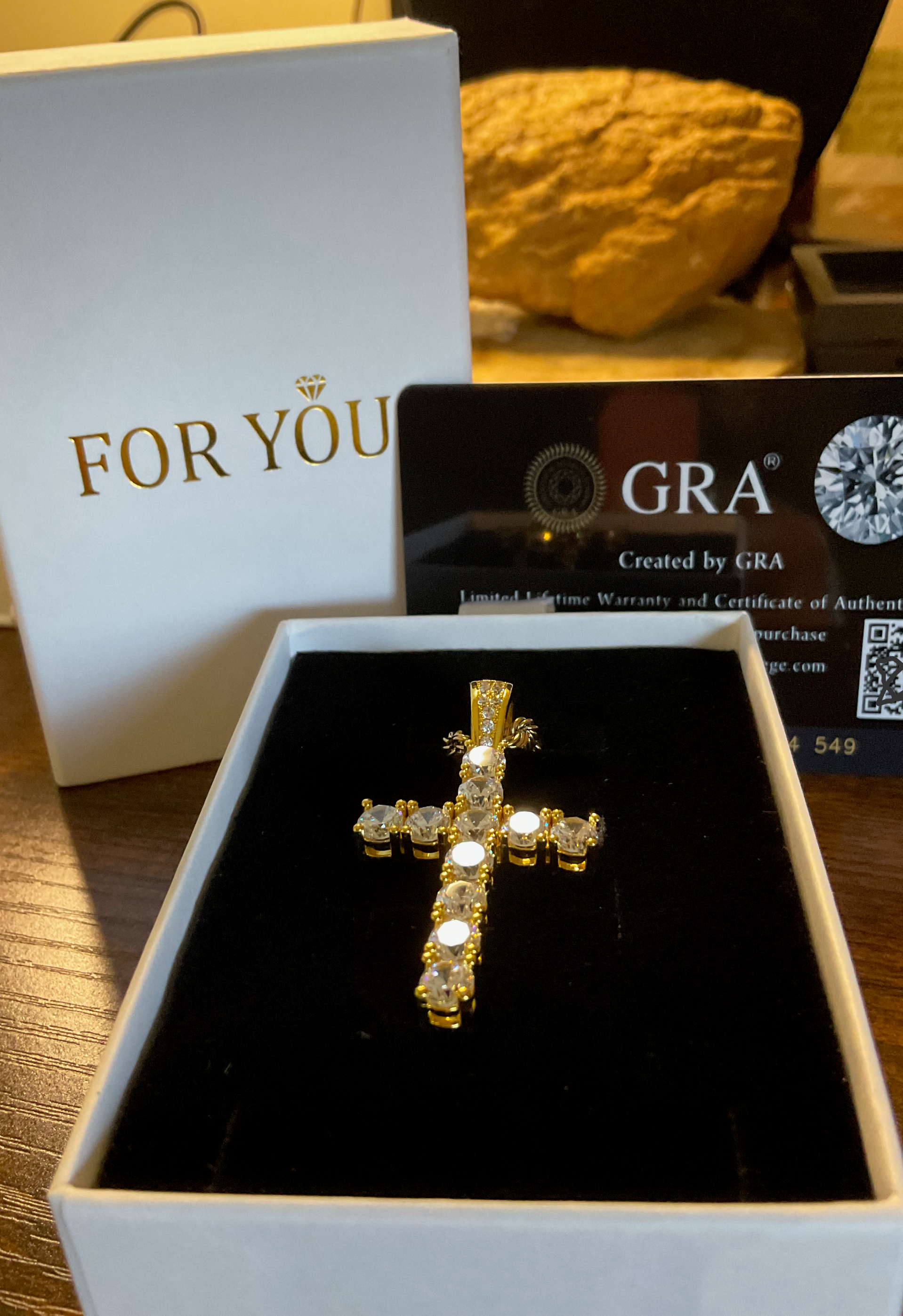 Gold cross pendant in a box with 'GRA' certification on a wooden surface