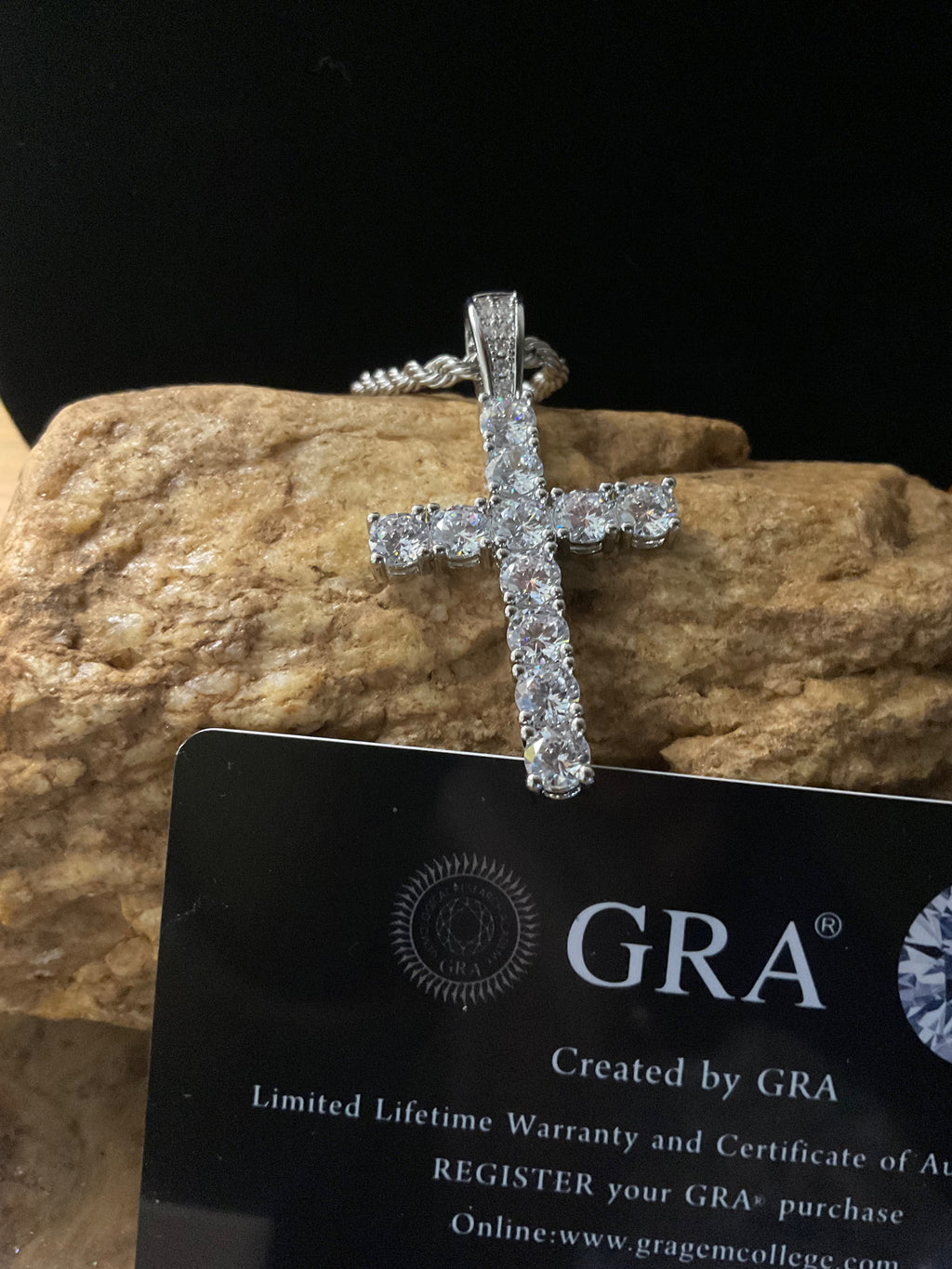 Diamond cross pendant on a black card with 'GRA' branding against a stone background