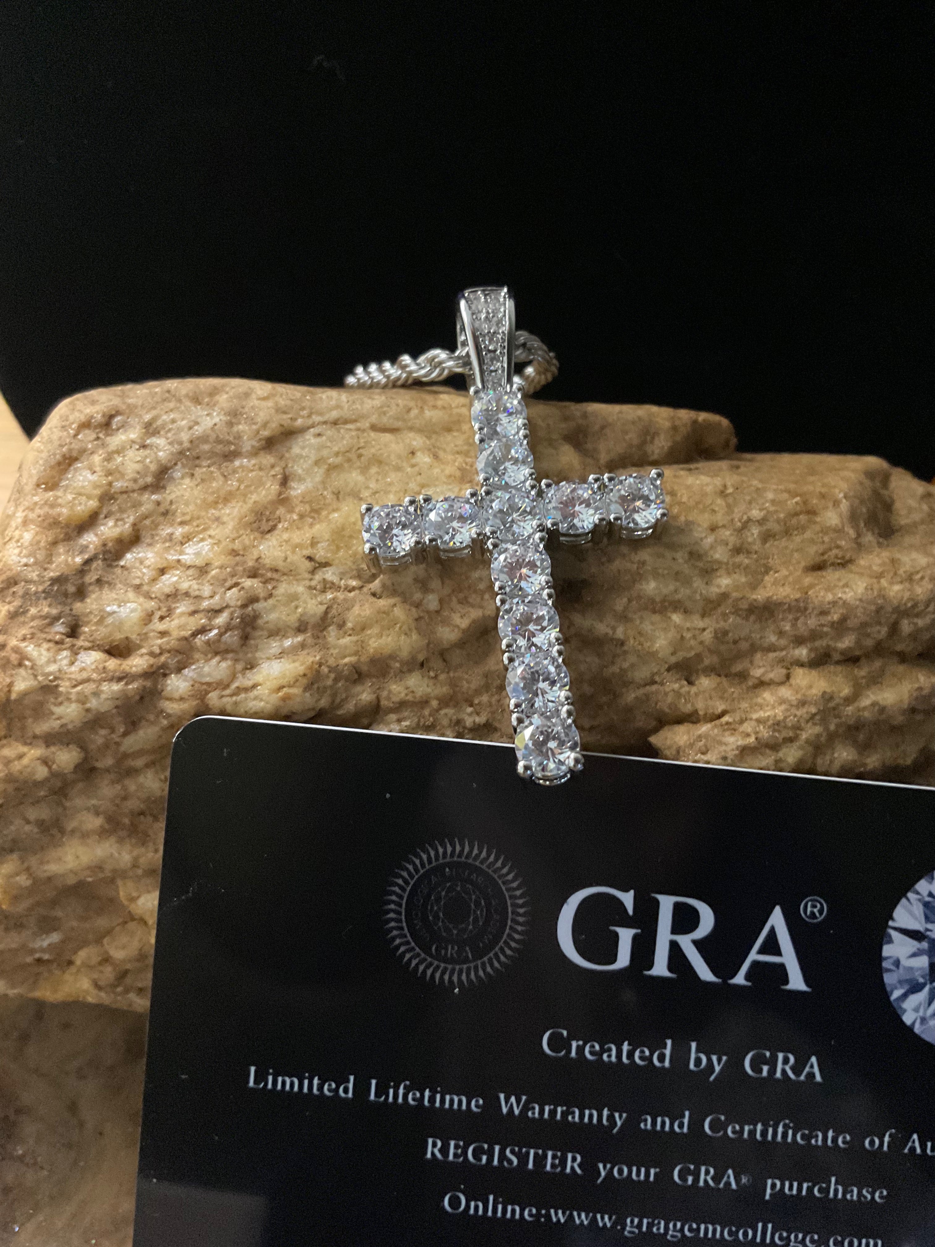 Diamond cross pendant on a black card with 'GRA' branding against a stone background