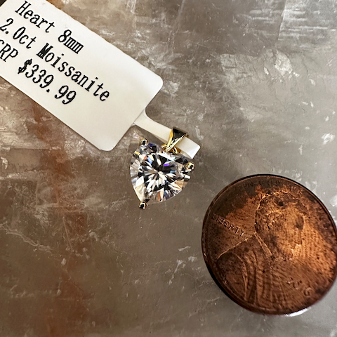 Heart-shaped moissanite gemstone with a price tag and coin for scale on a textured surface.