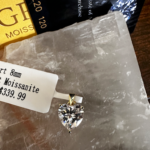 Heart-shaped moissanite pendant on a stone surface with a price tag and GIA certificate in the background.
