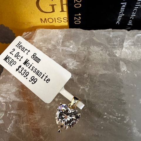 Gold pendant with a heart-shaped moissanite on a stone surface, with a tag displaying product details.