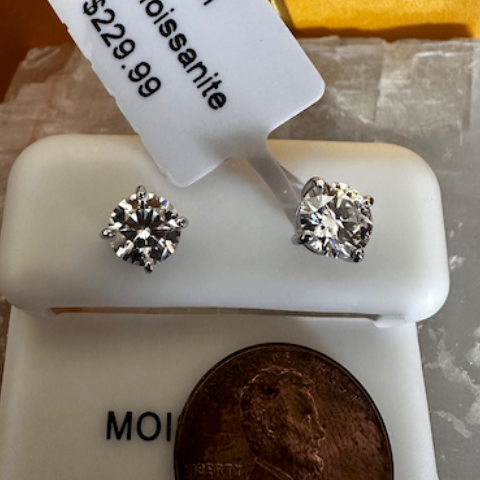 Round 6MM Moissanite earrings on a white stand with a penny for scale, against a gold and white background.