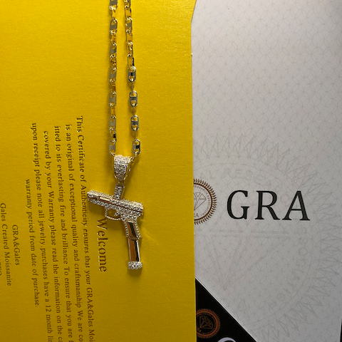 Gold necklace with a gun-shaped pendant on a yellow and white background with 'GRA' branding.