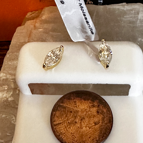Gold earrings on a white stand with a penny for scale, against a blurred background.