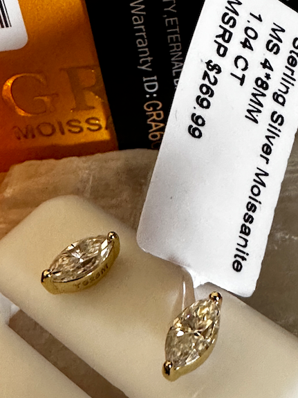 Gold earrings on a white stand with a visible brand label in the background.