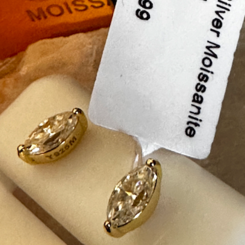Gold earrings on a white stand with a price tag displaying product details.