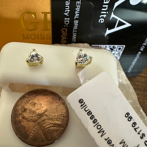 Gold earrings on a white stand with a coin for scale, against a blurred background.