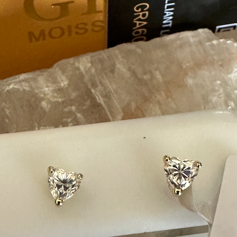 Heart-shaped diamond stud earrings on a white card with a gold box in the background.