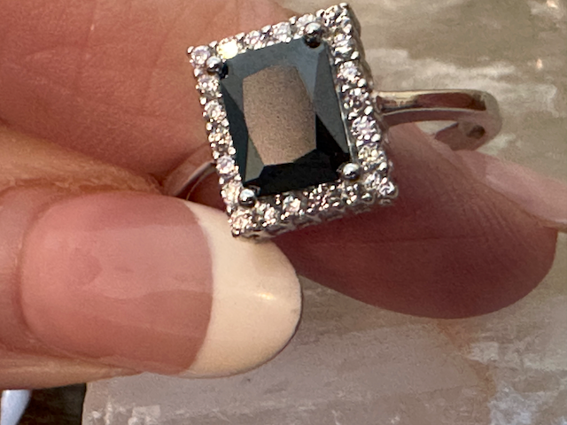 Ring with a black gemstone and diamond-studded square frame held between fingers.