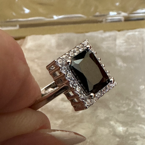Ring with a black gemstone and diamond-like stones held by a hand on a textured surface.
