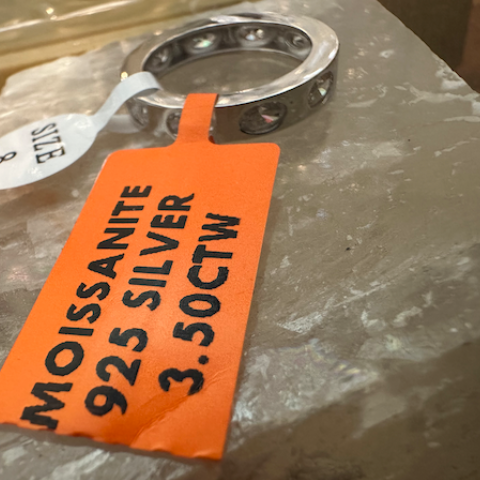 Silver ring with a Moissanite stone on a stone surface, accompanied by an orange tag with product details.