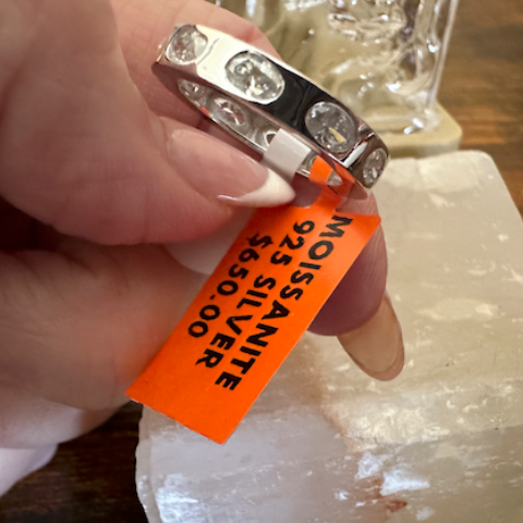Hand holding a silver ring with an orange tag on a wooden surface