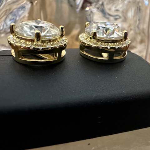 Gold earrings with clear gemstones on a black stand, blurred glass vase in the background.