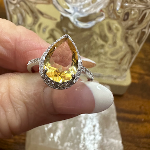 Teardrop-shaped yellow gemstone ring held by a hand with a decorative glass object in the background.