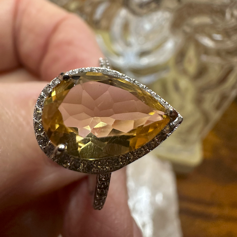 Ring with a large teardrop-shaped gemstone held by a hand, with a decorative object in the background.