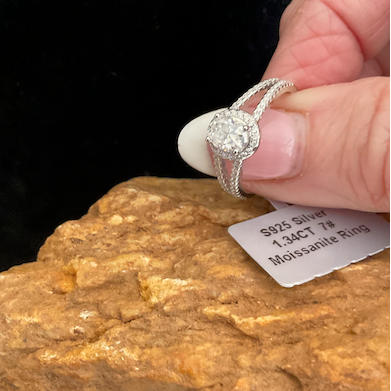 Diamond ring held by a hand above a rock with a label on a dark background
