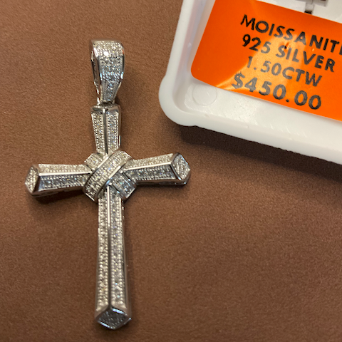 Silver cross pendant with moissanite stones on a brown surface, next to a price tag.