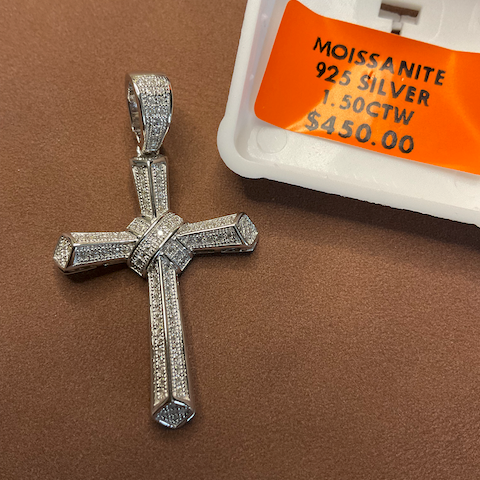 Silver cross pendant with moissanite stones on a brown surface, next to a price tag.