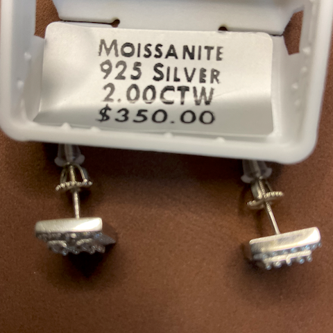 Silver earrings with moissanite stones on a white stand, showing product details.
