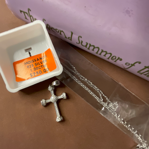 Silver cross necklace on a brown surface with a white box and orange label in the background.