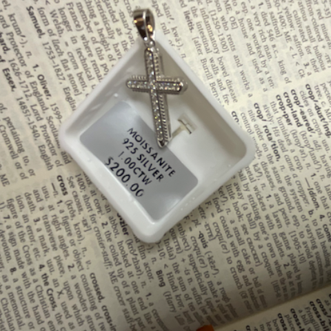 Silver cross pendant in a clear display case on a textured surface with text.