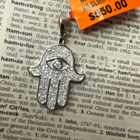 Silver Hamsa hand pendant with clear stones on a dictionary page with a price tag.