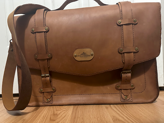 Brown leather satchel bag on a wooden floor with a white wall background